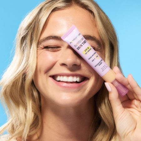 image of girl holding Revolution's SPF Skin Tint on a blue background